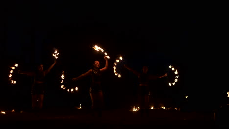 Three-women-with-burning-hoops-dance-with-fiery-torches-in-leather-clothes-in-a-dark-hangar-demonstrating-a-circus-fire-show-in-slow-motion